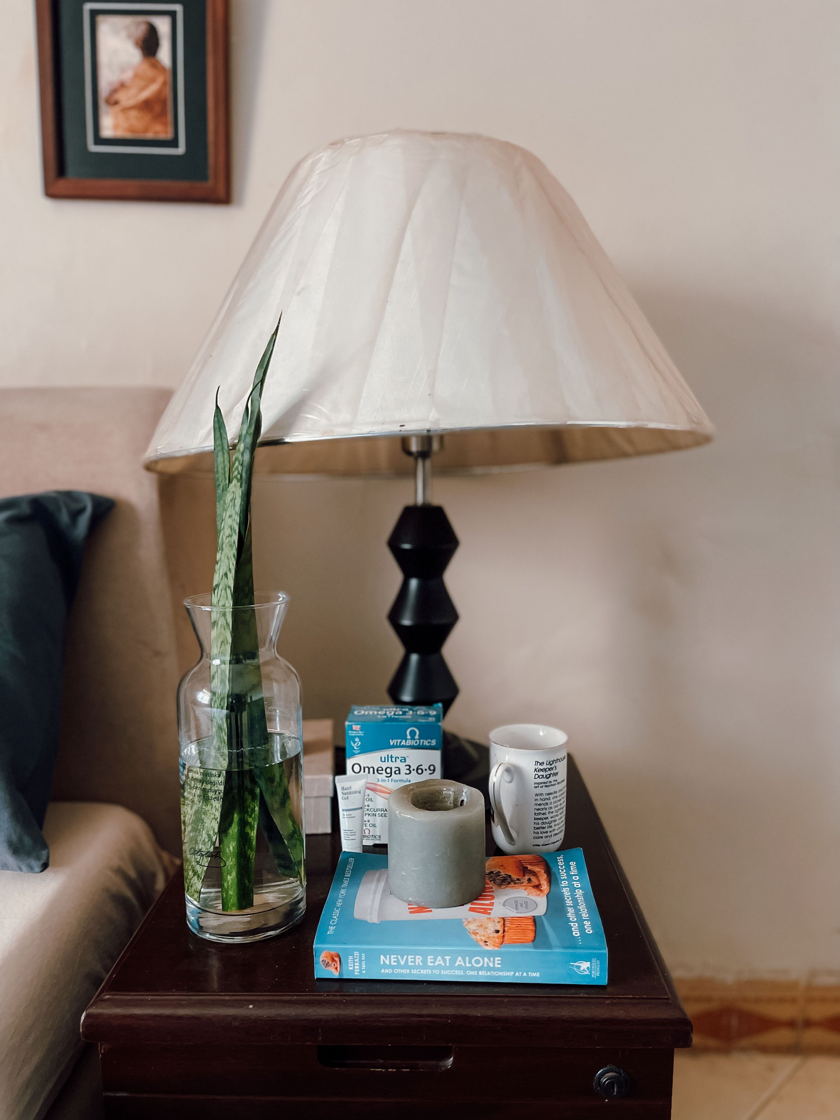 A bedside table with a lamp, a book titled 'NEVER EAT ALONE,' a box of Omega 3-6-9 supplements, a mug, and a glass vase with a plant.