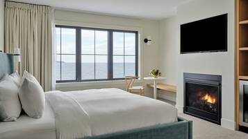 Quarto de hotel com uma cama grande, televisão na parede, lareira e vista para o mar através de uma janela ampla.