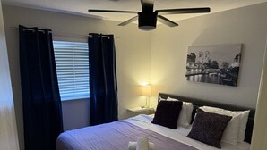 Habitació d'hotel amb llit, ventilador de sostre, tauleta de nit amb làmpada i un quadre a la paret.