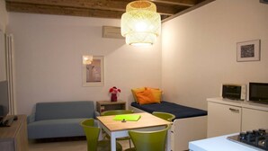 Una habitación compacta con cama, una mesa de comedor con sillas, kitchenette y una lámpara colgante.