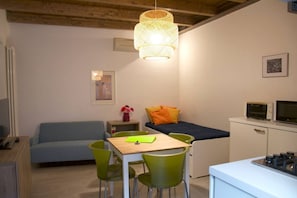 A compact room with a bed, a dining table with chairs, a kitchenette, and a hanging light fixture.