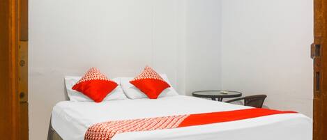 A hotel room with a bed, two red pillows, a white bedspread, and a small round table with a chair.