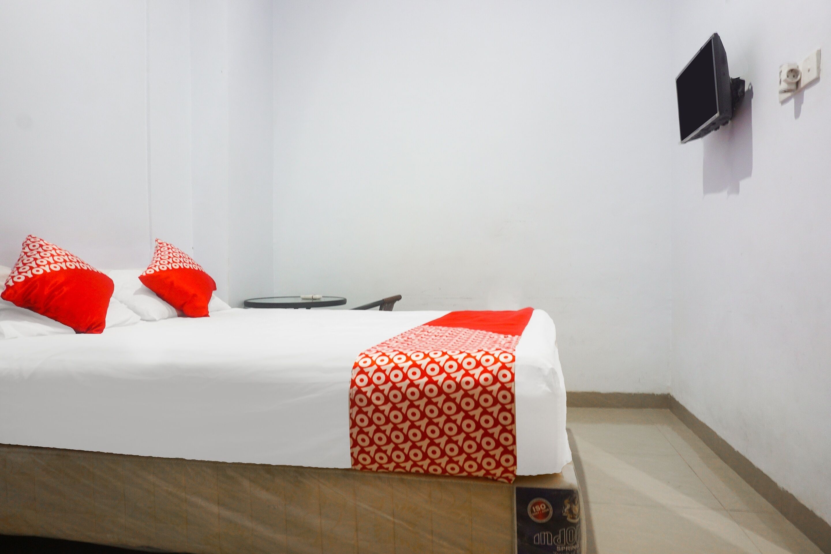 A single bed with white bedding and red pillows, a bedside table with a black object, and a wall-mounted TV.