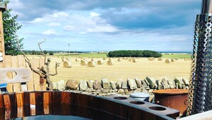 A hot tub with a stone wall and wooden deck, overlooking a field of hay bales and a distant forest. - The Sanderling  - sleeps 2 guests  in 1 bedroom (Beadnell)