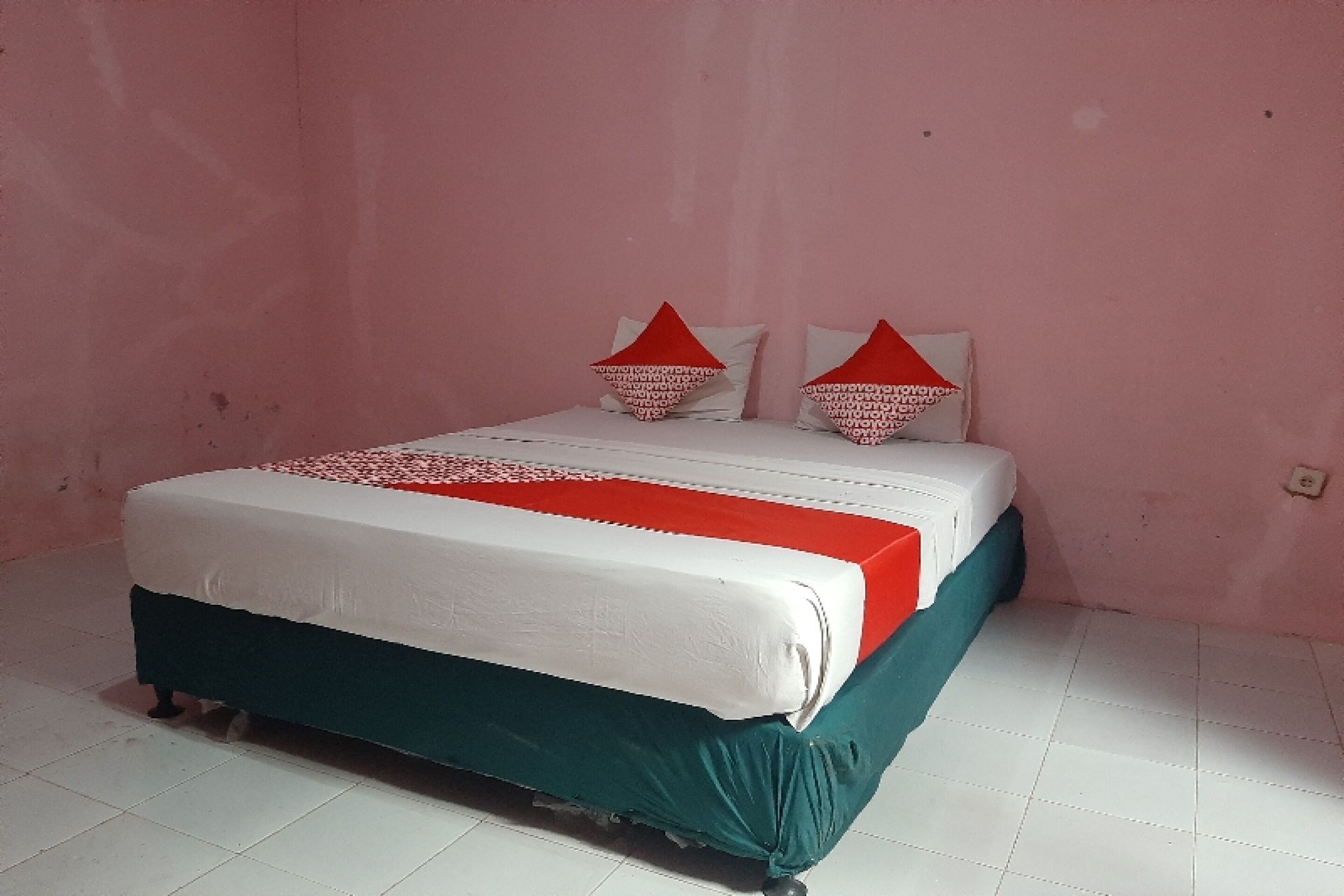 Un lit recouvert d’une couvre-lit blanche et rouge, avec une jupe de lit turquoise et des oreillers rouges et blancs, dans une pièce aux murs roses et au sol carrelé de blanc.