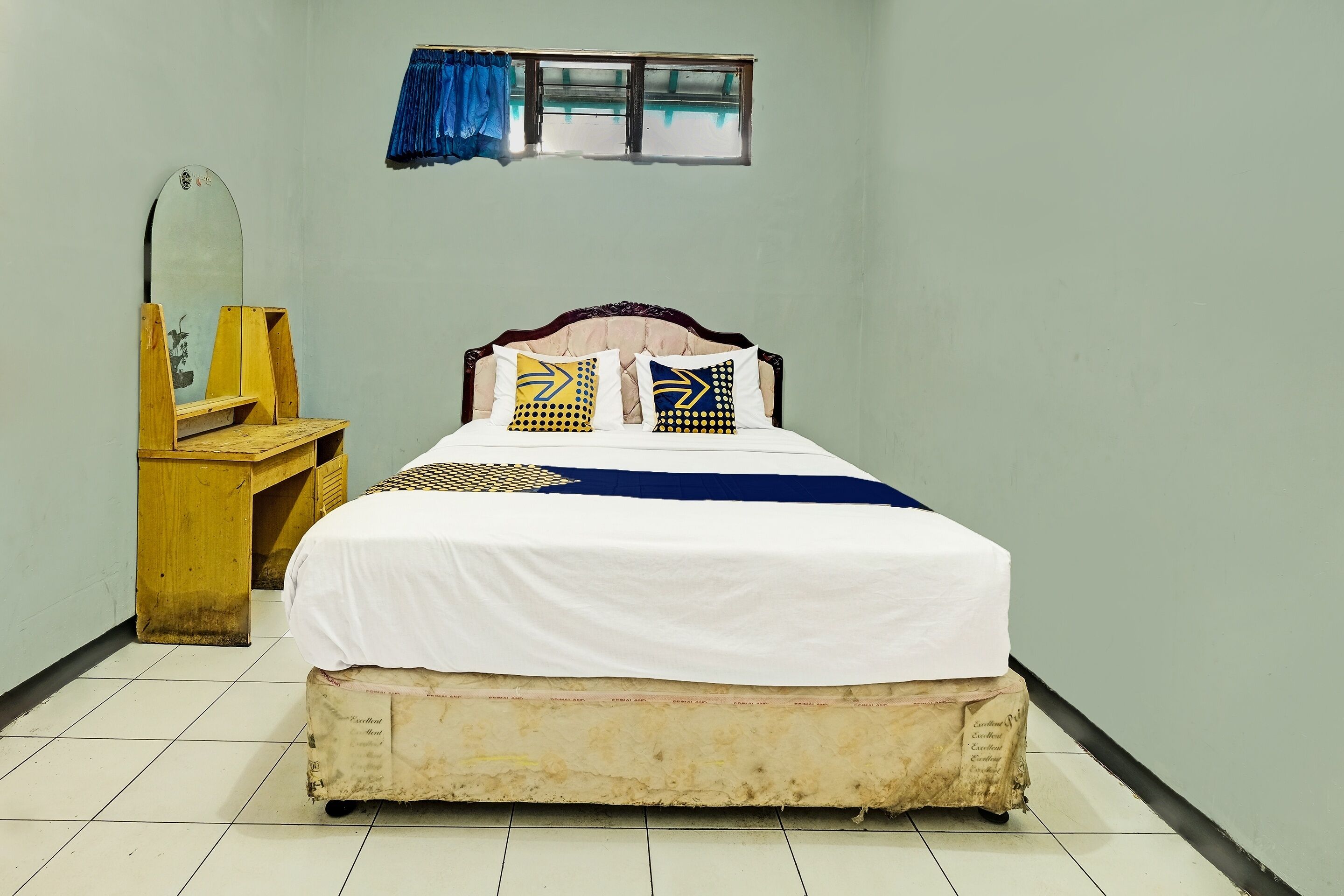 A single bed with a white bedspread and patterned pillows, a wooden nightstand, a mirror, and a window with blue curtains.