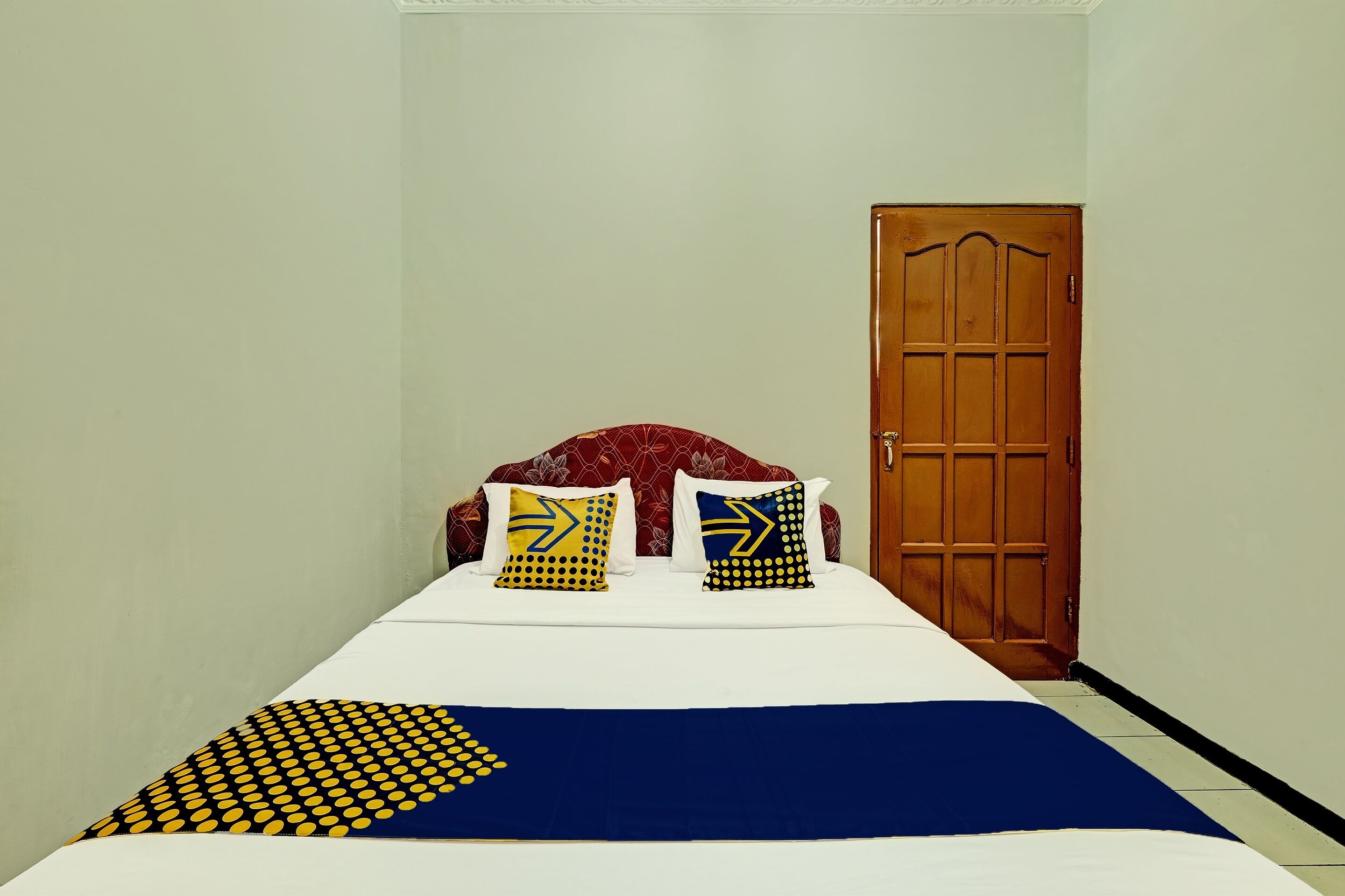 A single bed with a patterned bedspread and two pillows with geometric designs. A wooden door is visible to the right.