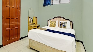 A bedroom with a wooden door, a bed with patterned pillows, a small wooden nightstand, and a window with blue curtains. - SPOT ON 91715 Hotel Citra Dewi 1 (Bandungan)