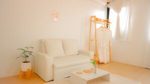 A minimalist living room with a white sofa, a small coffee table with decorative items, a wooden chair, a plant, and a clothing rack. - HaneulHotelChiamai (Chiang Mai)