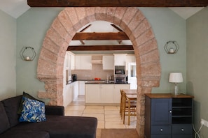 A living room with a sofa, a coffee table, and a TV. There is a kitchen visible through an archway with white cabinets and a microwave.