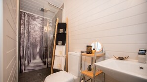 Un baño moderno con inodoro, estante de madera con artículos de aseo, espejo y cabina de ducha de vidrio con vista al bosque.