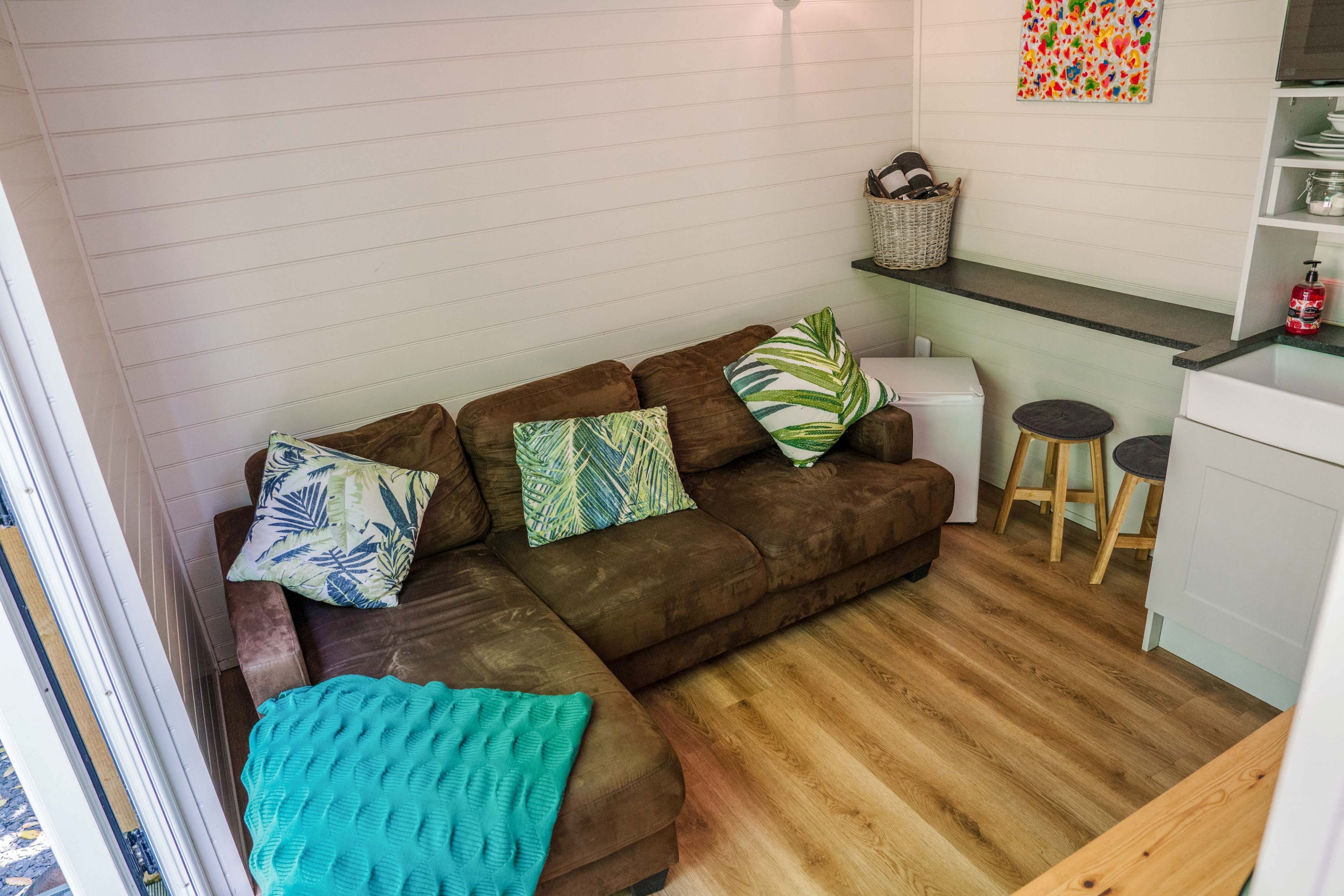Una sala de estar acogedora con un sofá marrón, una manta color esmeralda y una cocina con taburetes y un refrigerador blanco.