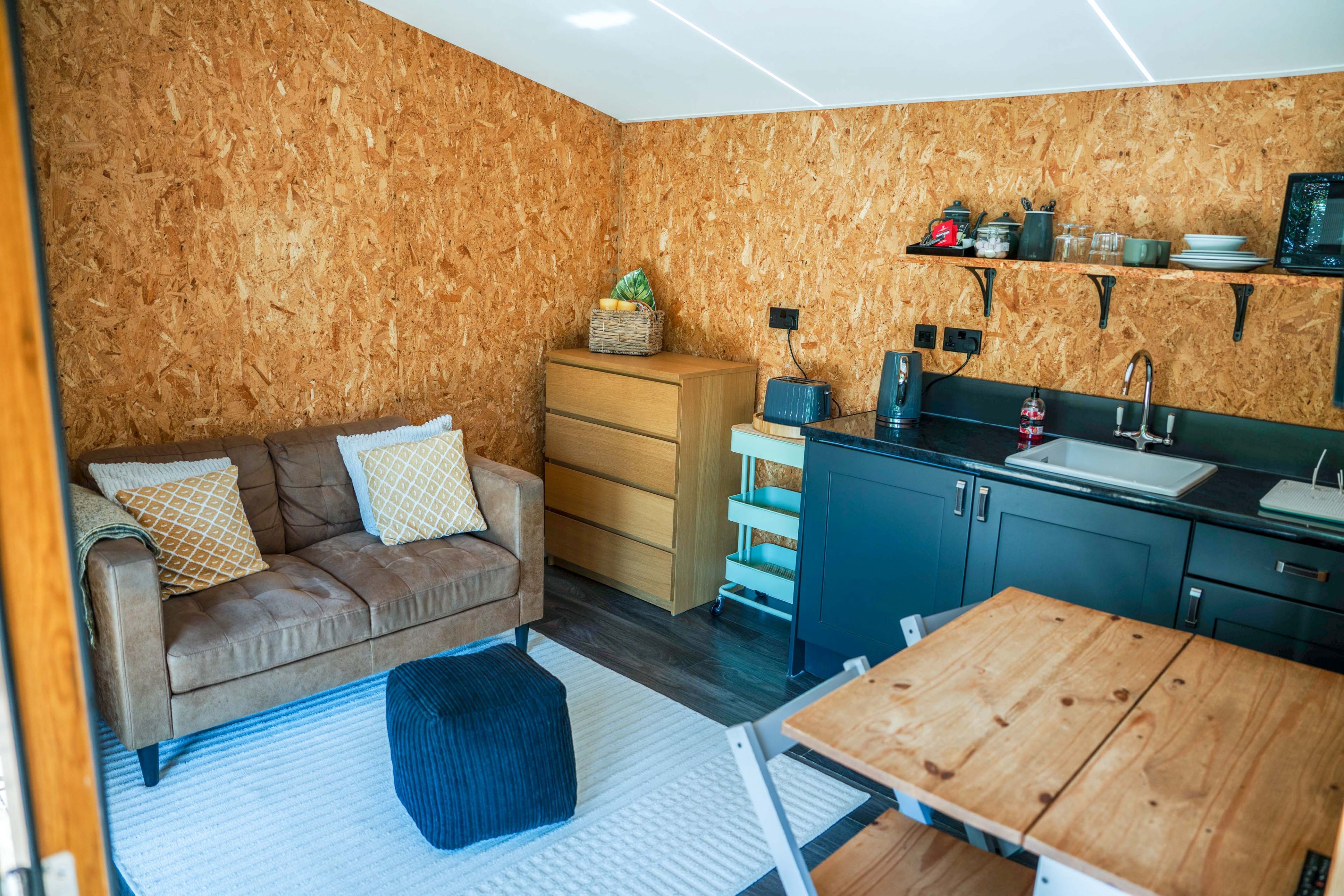 Un espace de vie compact comprenant un coin cuisine, un canapé, une table en bois et un petit tapis.