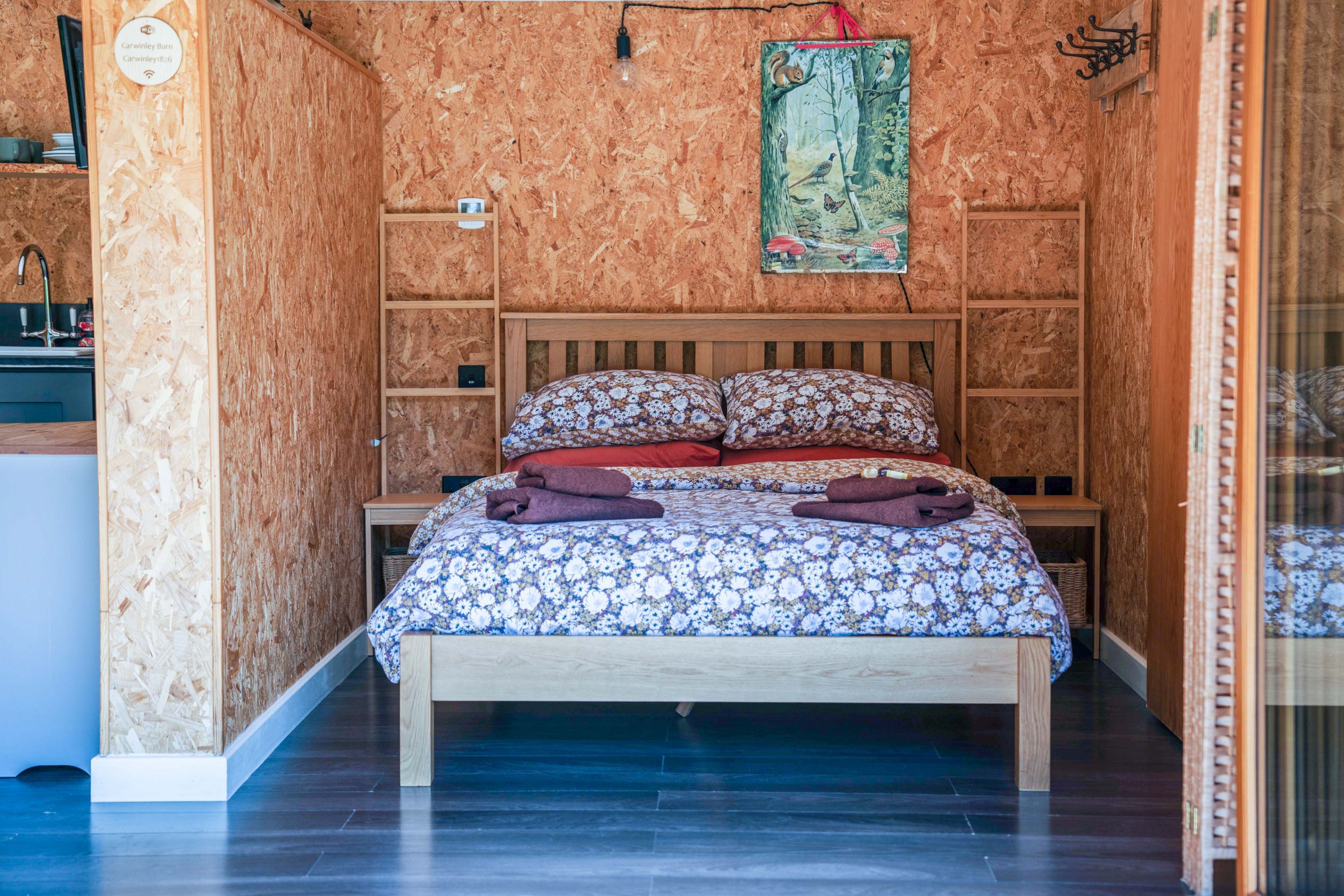 Ein Schlafzimmer mit einem hölzernen Kopfteil, einem Bett mit floraler Bettwäsche, einem kleinen Nachttisch, einem Wandregal und einem Bild an der Wand.