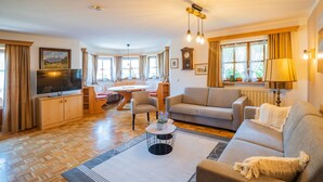 A spacious living room with a sofa, a coffee table, and a TV. There is a dining area with a table and chairs. The room has wooden flooring and large windows with curtains.