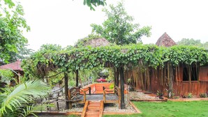 A wooden bridge over a garden with a thatched roof structure, surrounded by greenery and a red car in the background. - OYO 92083 Lailik Gilang Guesthouse (Werinama)