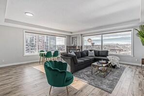 Un salón moderno con un sofá modular, sillones verdes y una mesa de centro. Amplias ventanas ofrecen vistas a un paisaje urbano.