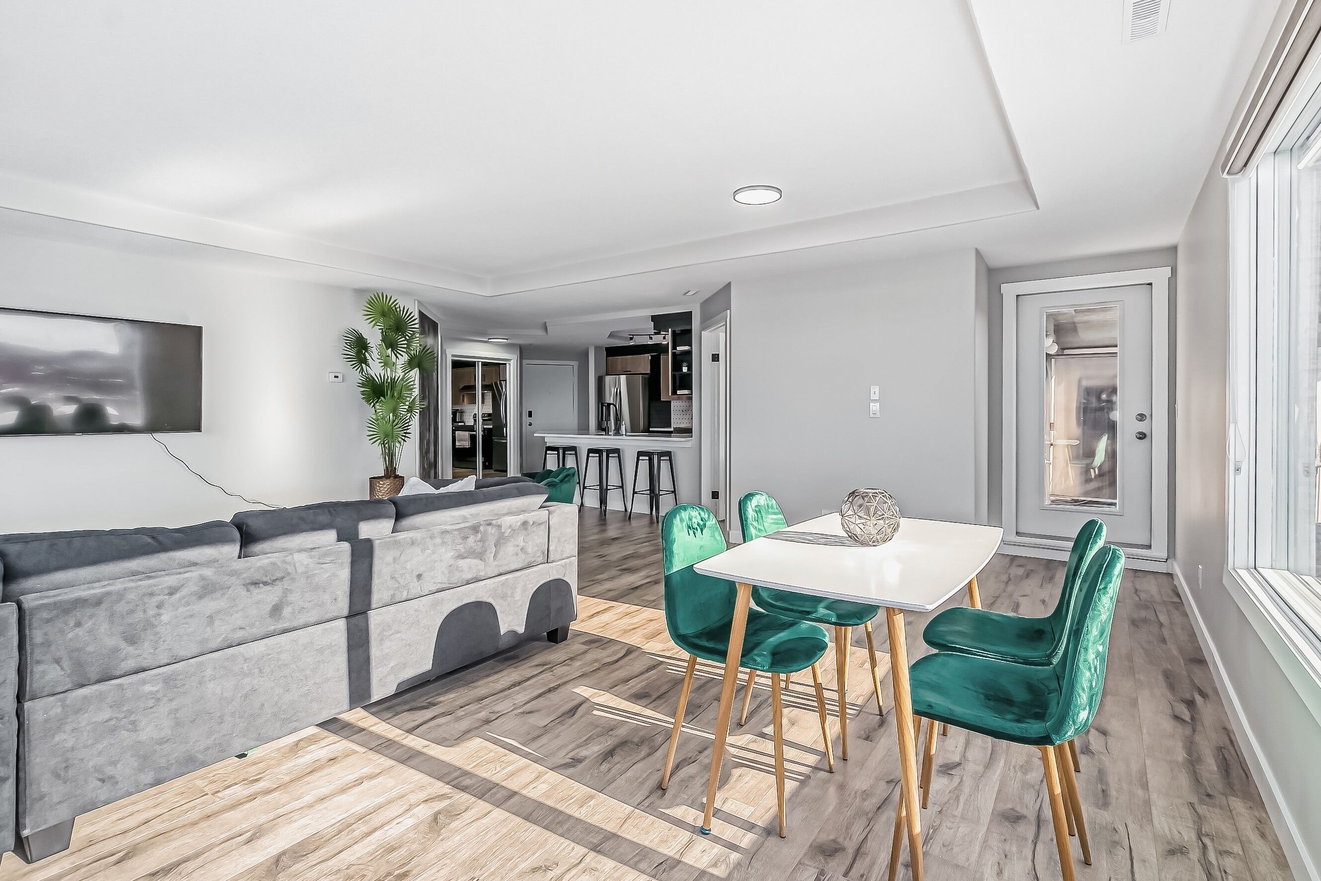 Un salon moderne avec un canapé gris, une table à manger blanche et des chaises vertes, ainsi qu’un téléviseur à écran plat fixé au mur.