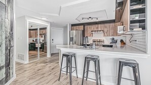 Een moderne keuken met een wit aanrecht, roestvrijstalen apparatuur en houten kastjes. Er staan drie krukken bij het aanrecht en een schuifdeur die naar een andere kamer leidt.
