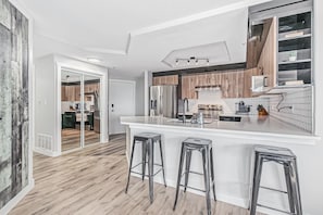 Cozinha moderna com bancada branca, eletrodomésticos de aço inoxidável e armários de madeira. Há três banquetas no balcão e uma porta deslizante que dá para outro cômodo.