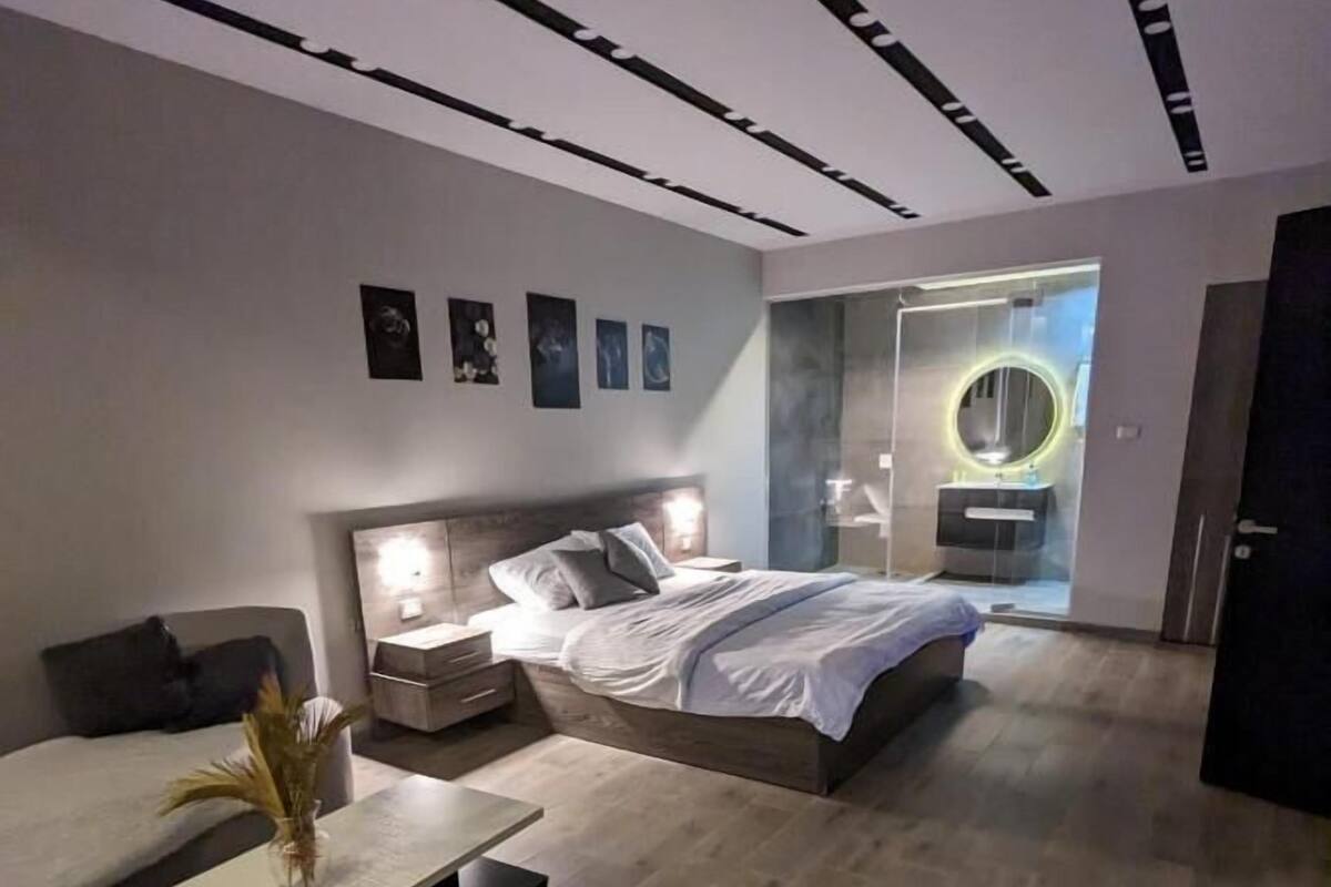 A modern bedroom with a bed, bedside tables, a nightstand, a small table with a vase, and a wall-mounted circular mirror.