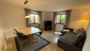 Een moderne woonkamer met een houten eettafel, twee stoelen, een grijze bank, een klein witte bijzettafel en een tv-meubel.