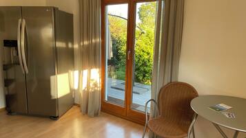 Eine moderne Küche mit einem Edelstahlkühlschrank, eine Essecke mit einem Korbstuhl und einem kleinen Tisch sowie eine Glastür, die auf eine Terrasse mit Grünfläche im Freien führt.