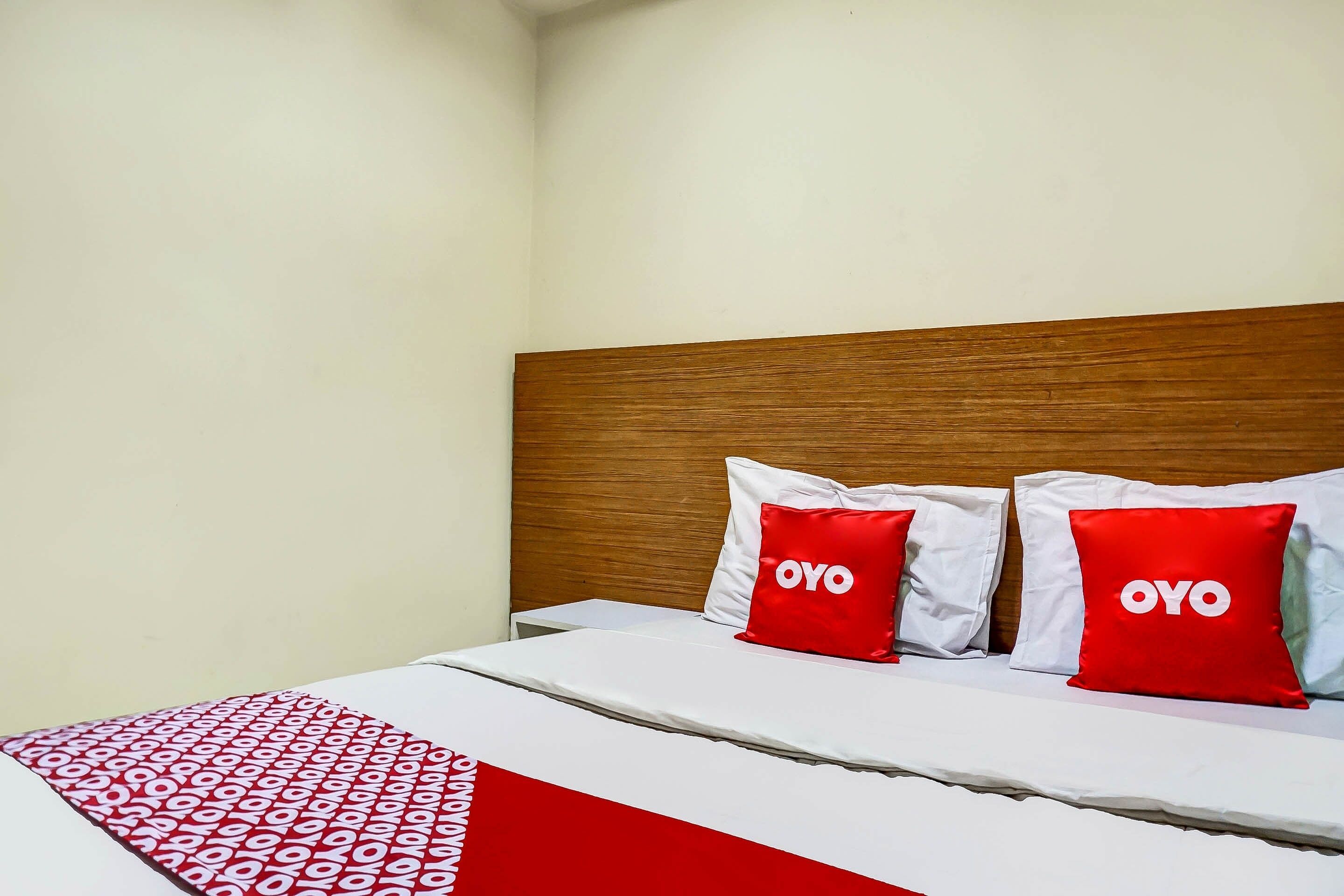 Una habitación de hotel con cabecera de madera, cama con ropa blanca y almohadas rojas, y una manta roja y blanca estampada.