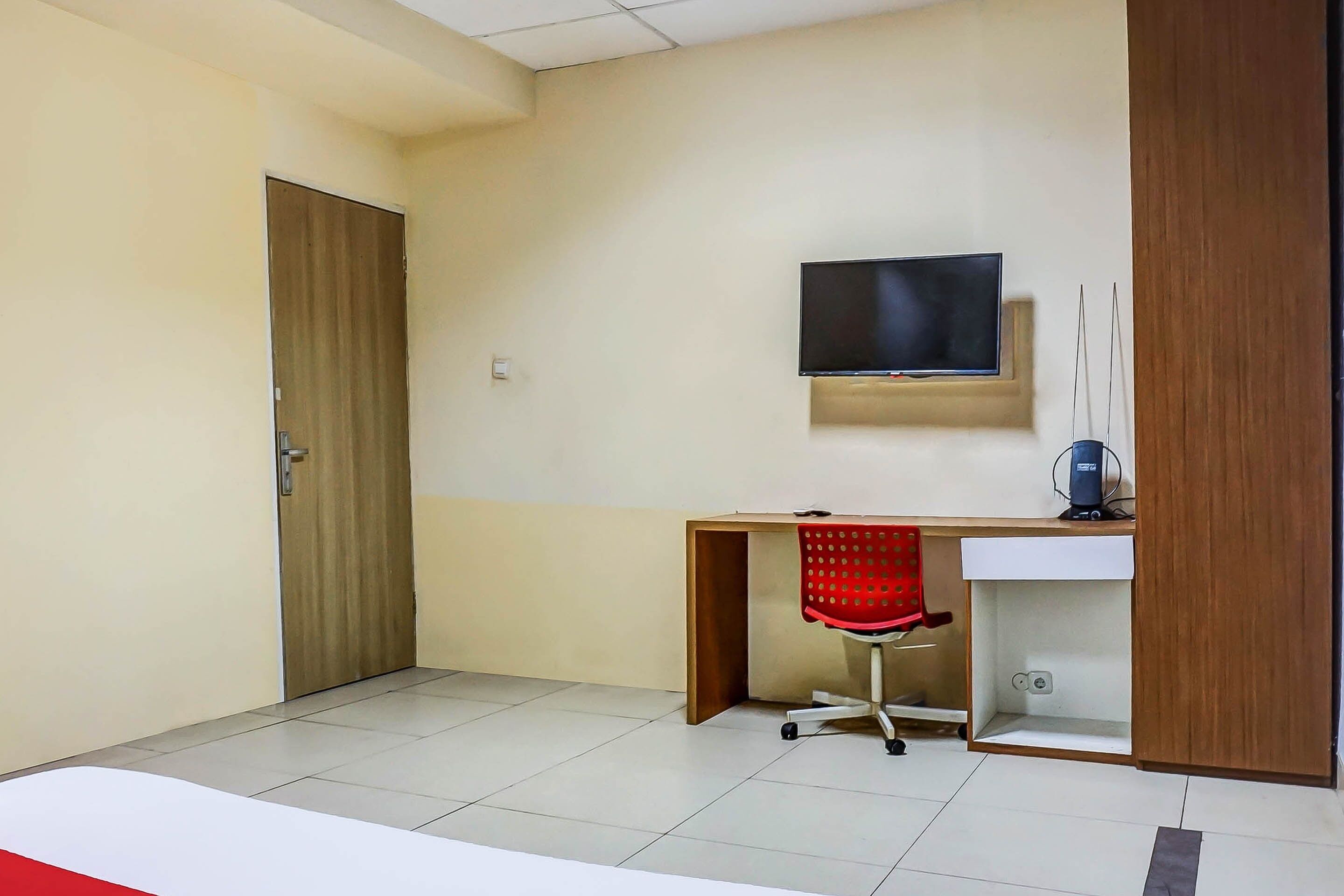 Une pièce avec une porte en bois, un bureau avec un fauteuil de bureau rouge, un téléviseur fixé au mur et un lavabo blanc.
