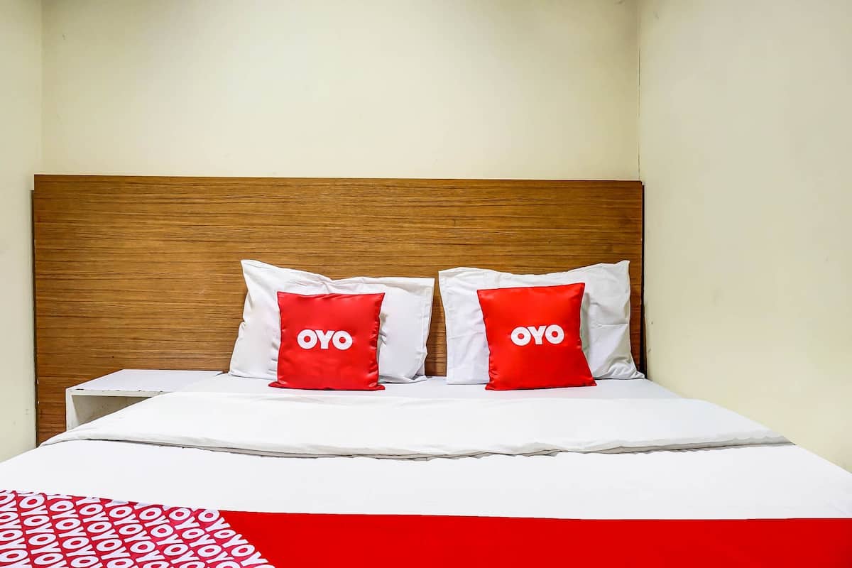 Une chambre d’hôtel avec une tête de lit en bois, du linge de lit blanc et deux oreillers rouges ornés du logo « OYO ».