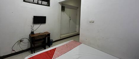 A small, sparsely furnished room with a bed, a desk, a television mounted on the wall, and a door leading to another room.