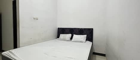Quarto pequeno com cama de solteiro, um edredom branco simples e duas almofadas. A cabeceira tem um padrão floral, e há uma pequena porta escura no lado esquerdo.