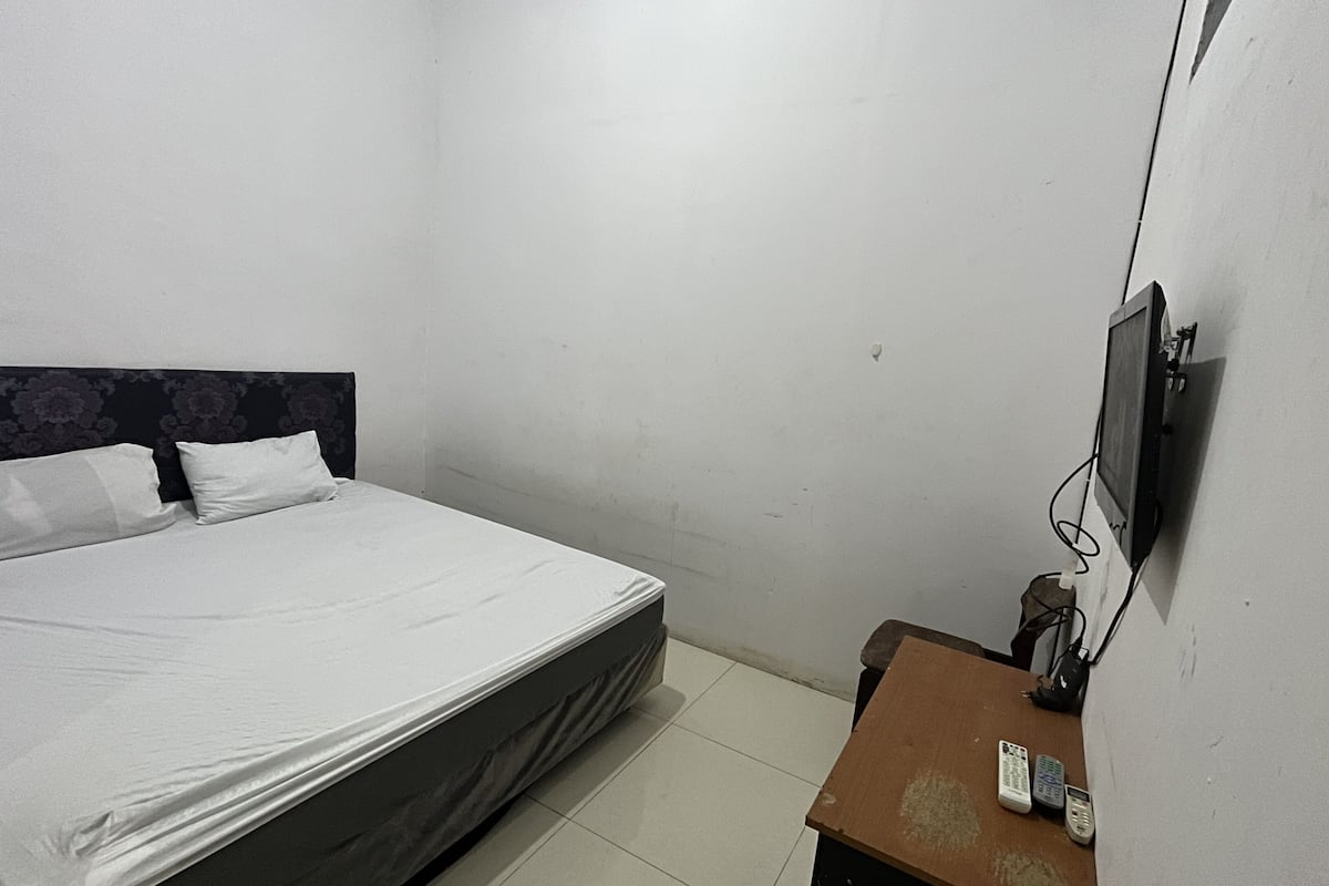 A single bed with a headboard, a flat-screen TV mounted on the wall, and a small wooden nightstand with two remote controls.