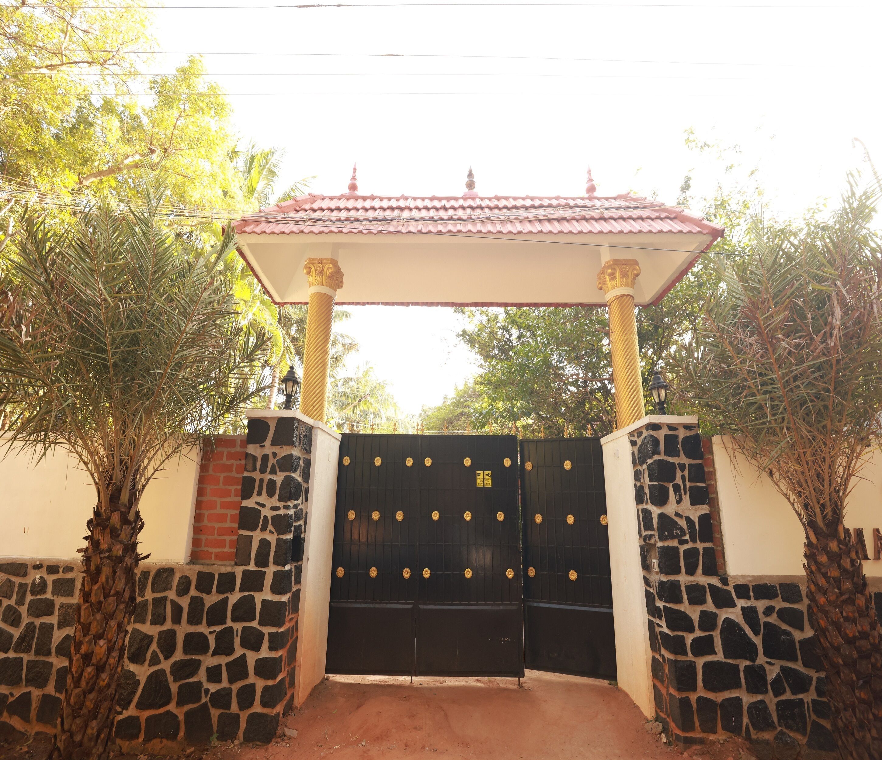 Una entrada con reja, techo de tejas, muros de piedra y palmeras.