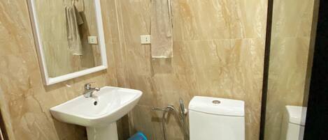 Une salle de bain avec un lavabo, des toilettes et un miroir. On y trouve également des plantes et une poubelle bleue.