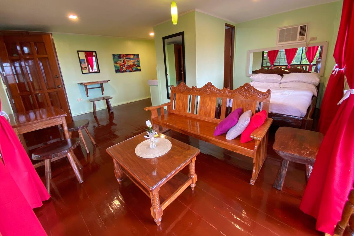 A room with a wooden bench, a small table with a vase, a bed with red curtains, and a painting on the wall.