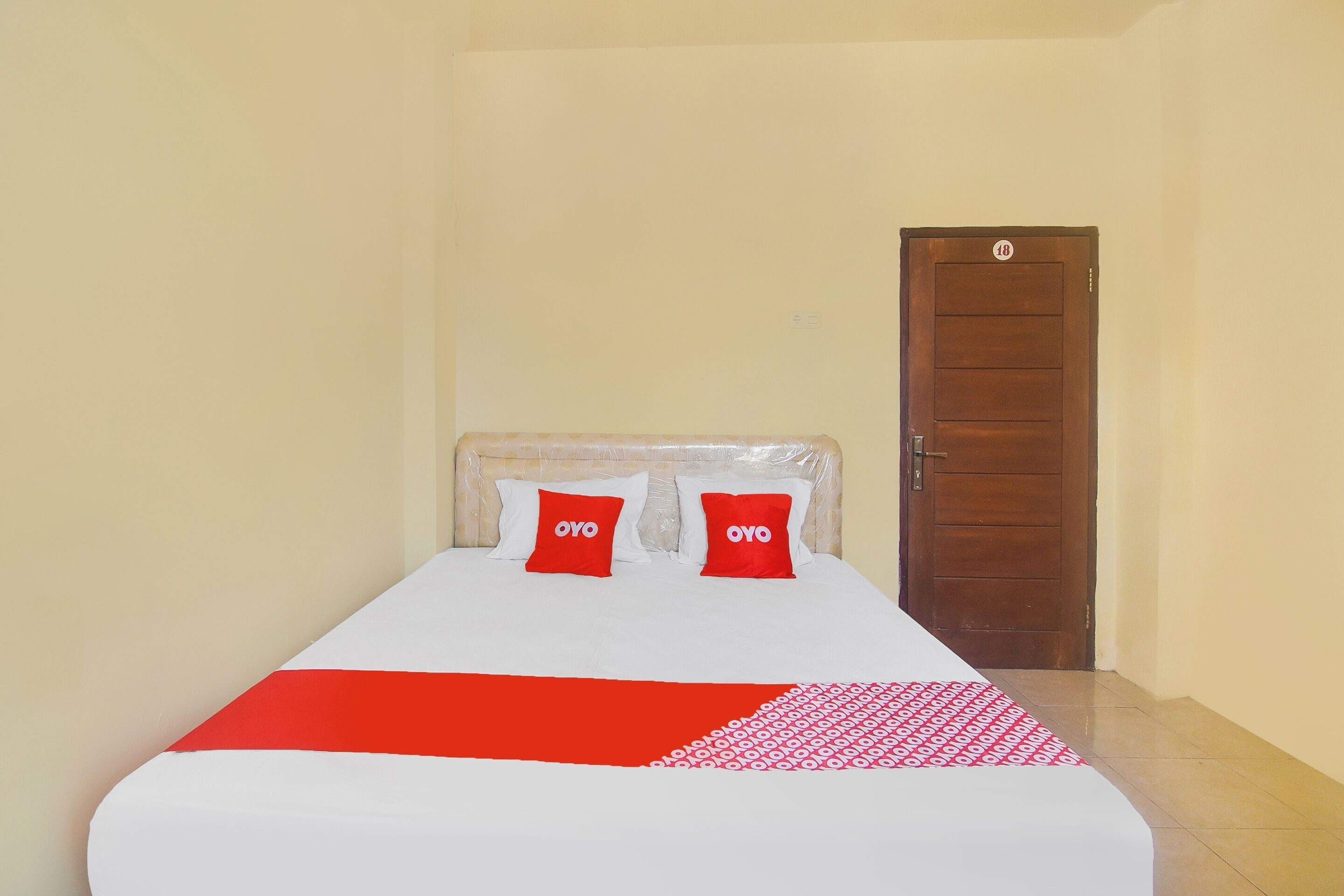 A single bed with a white and red bedspread, two red pillows with the word "OYO" on them, and a wooden door in the background.
