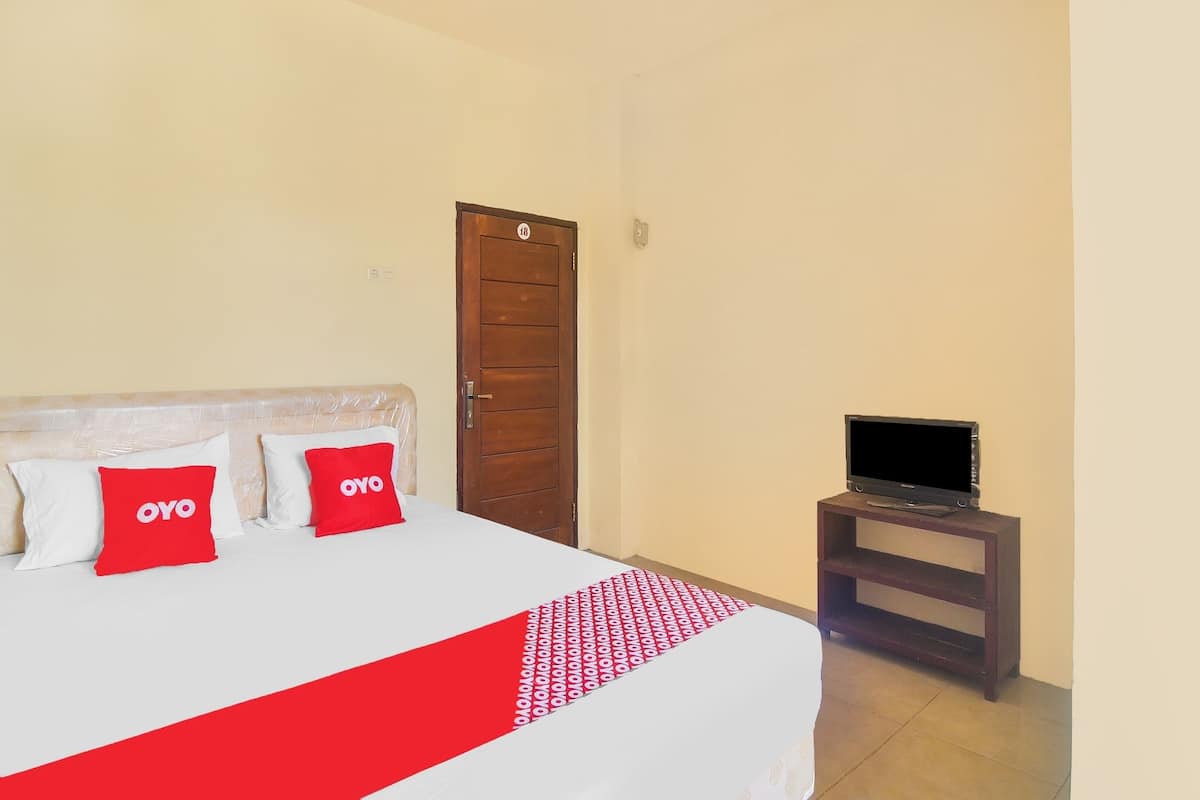 Quarto de hotel com uma cama, duas almofadas vermelhas com a marca 'OYO', uma televisão sobre um suporte de madeira e uma porta com fechadura.