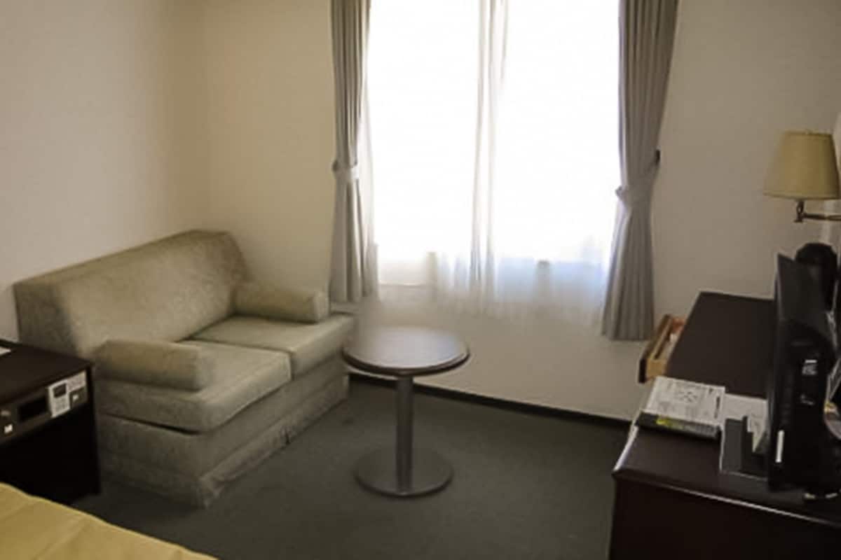 Una habitación de hotel con una cama, un sofá, una mesita redonda, un escritorio con lámpara y una ventana con cortinas.