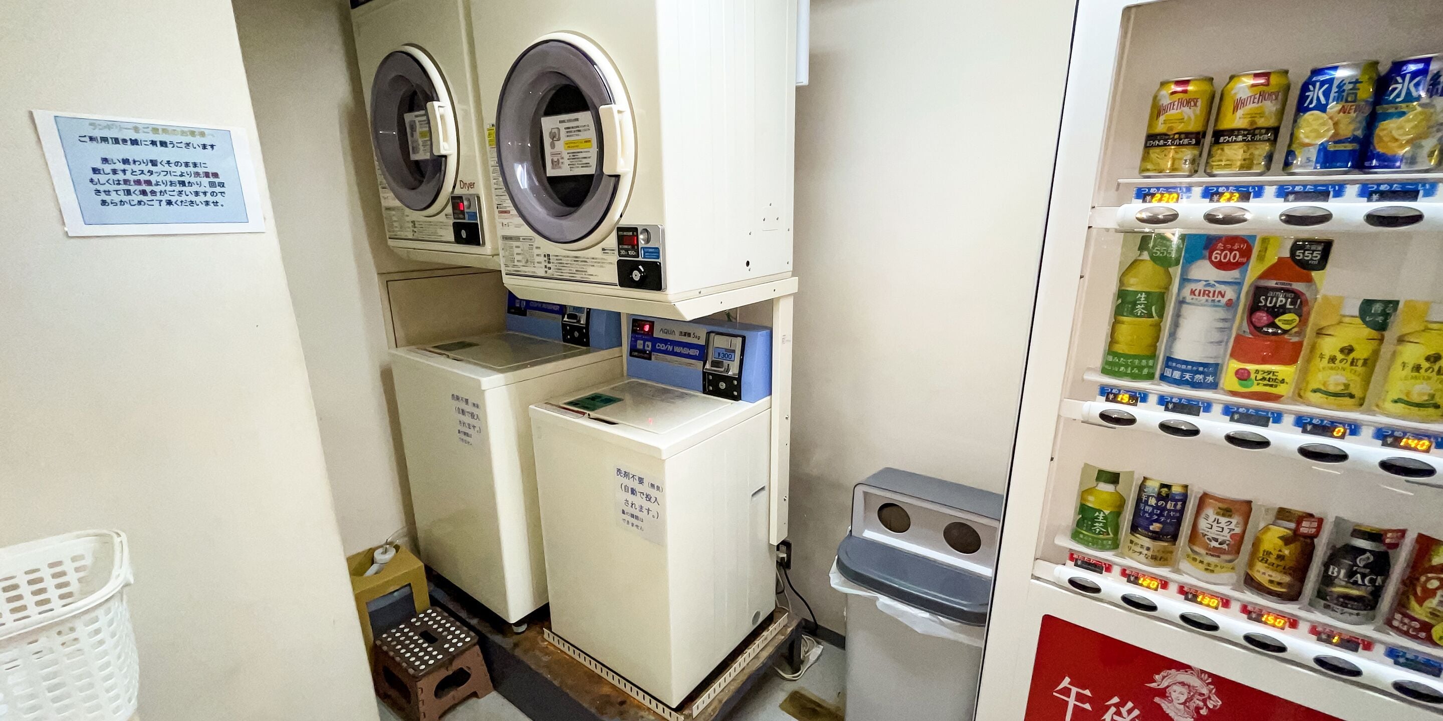 A laundry area with two washing machines and a dryer, a small stool, and a shelf with various beverages.