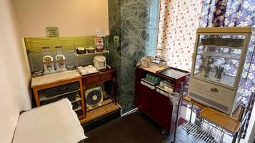 A small kitchenette with a coffee maker, a fan, and a microwave. There is a small shelf with condiments and a basket.