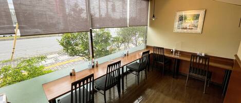 A restaurant with a long wooden table and black chairs, large windows with blinds, and a framed picture on the wall.