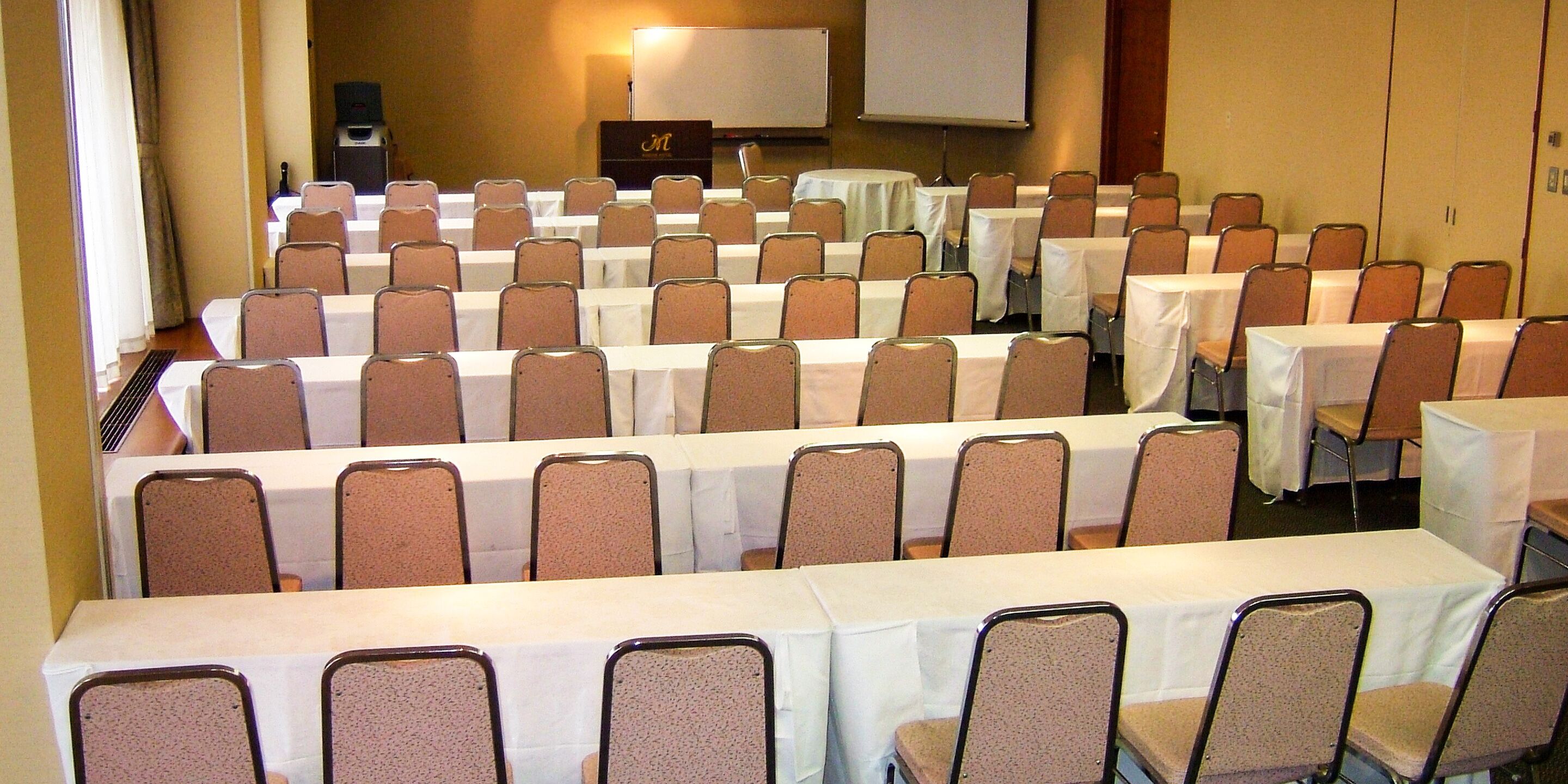 A conference room with rows of tables and chairs, a whiteboard, and a projector screen.