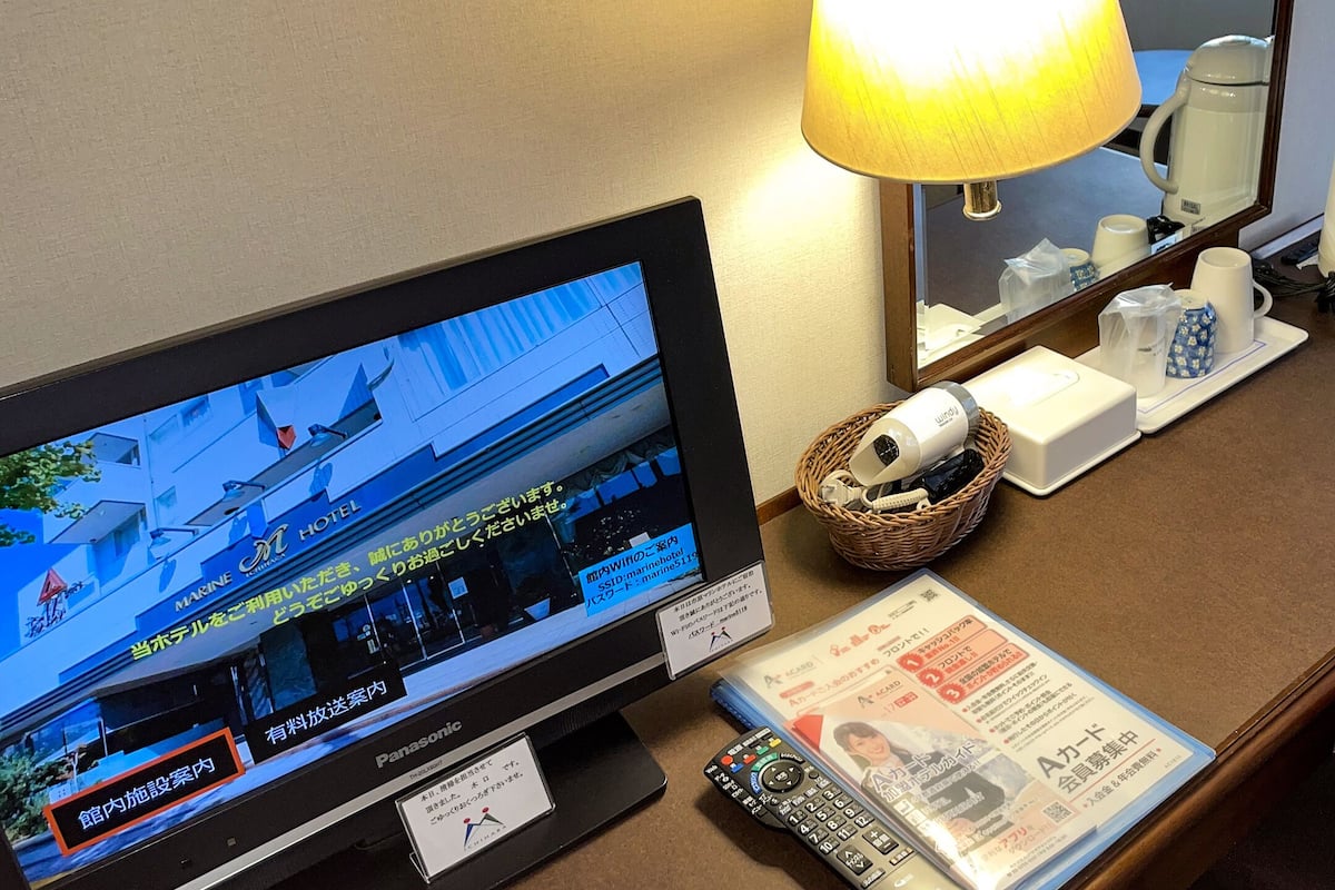 Habitación de hotel con una televisión mostrando el exterior de un edificio, una lámpara, una cafetera, un control remoto y un folleto sobre la mesa.