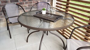 Une table ronde en verre avec un cadre en métal, accompagnée de deux chaises et d’un plateau sur lequel est inscrit le texte : « À TABLE ! ».