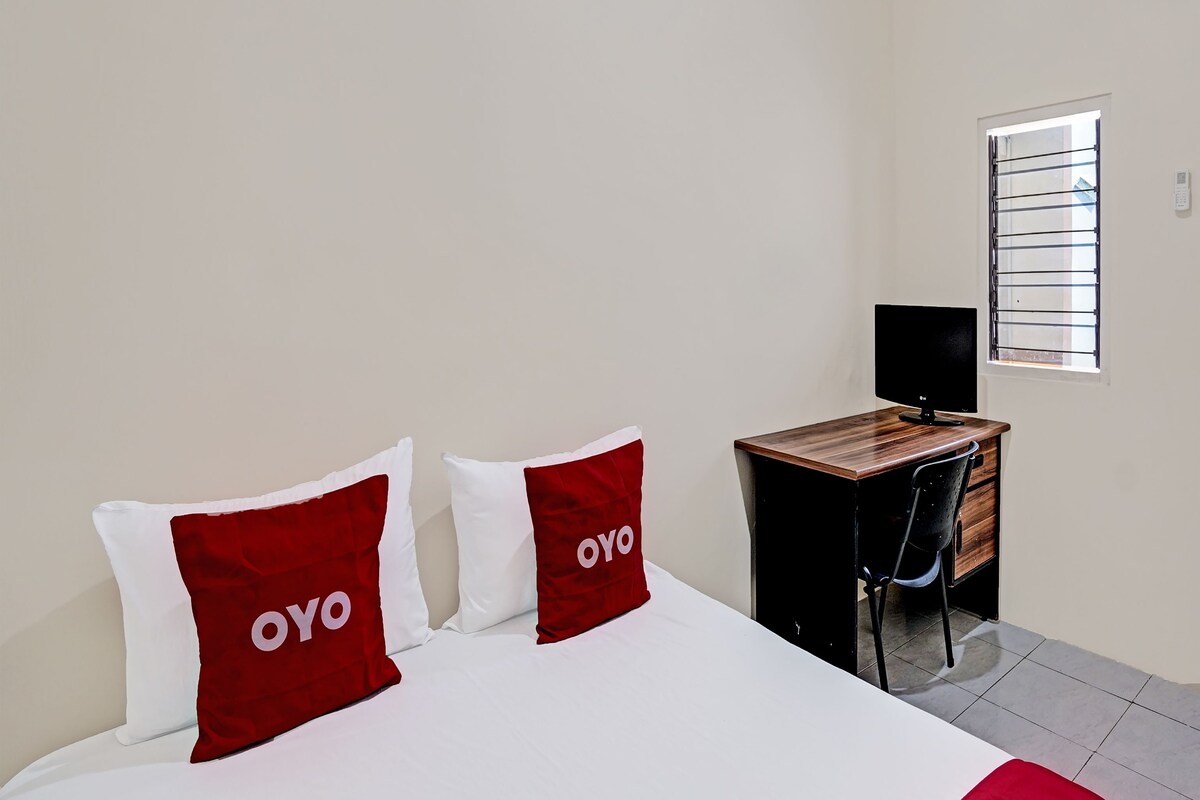 Ein Hotelzimmer mit einem Bett, zwei Kissen mit "OYO" Branding, einem Schreibtisch mit einem Computer und einem Fenster mit Jalousien.