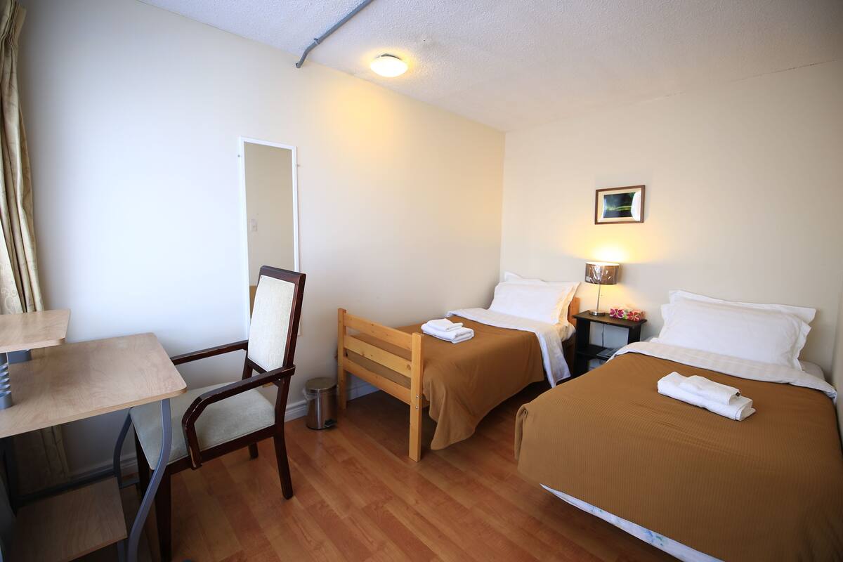 Une chambre d’hôtel avec un bureau en bois, deux lits simples, une chaise, une table de chevet et une petite poubelle.