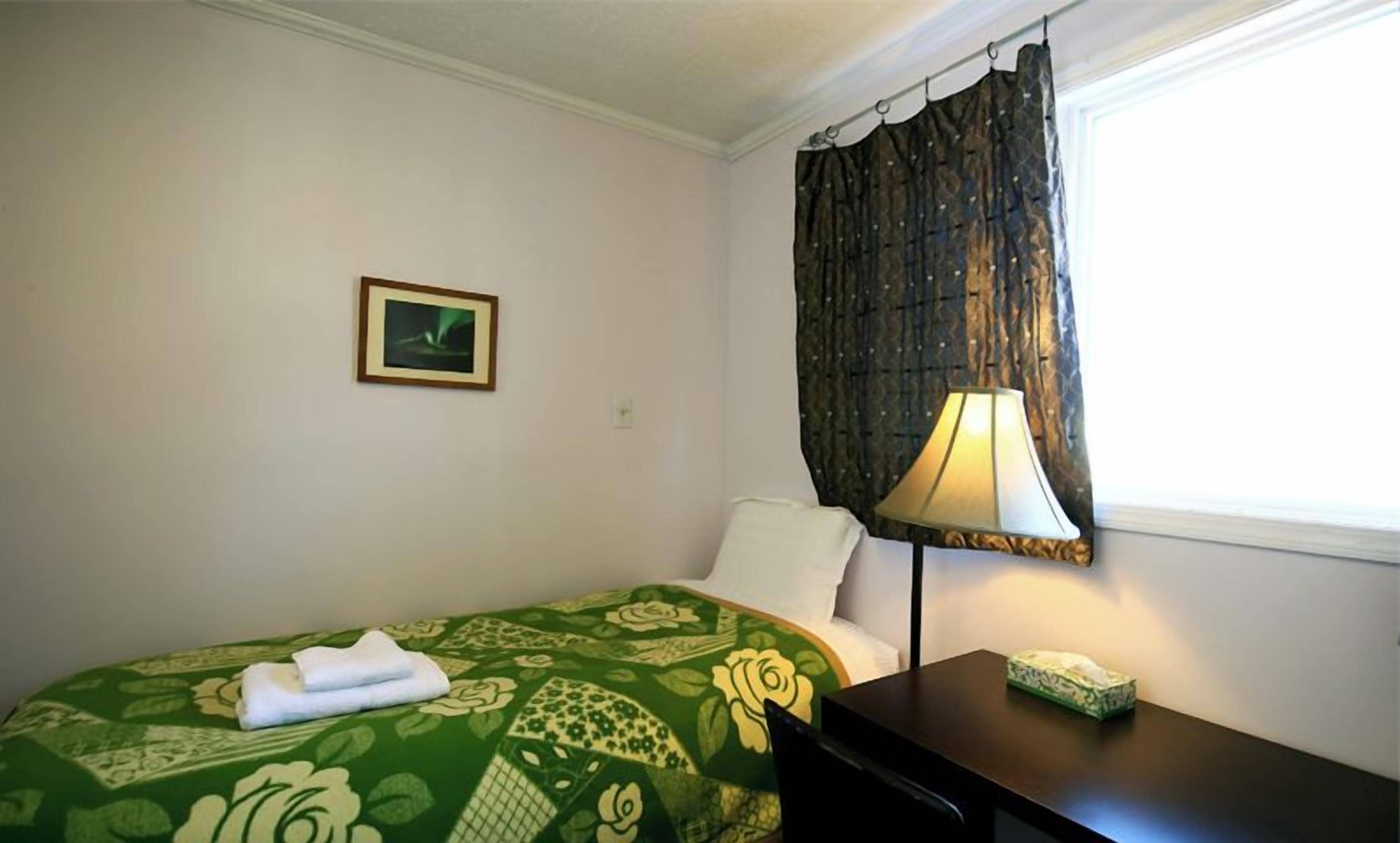 A hotel room with a green floral bedspread, a white pillow, a framed picture on the wall, a dark wooden desk with a lamp, and a window with a dark curtain.