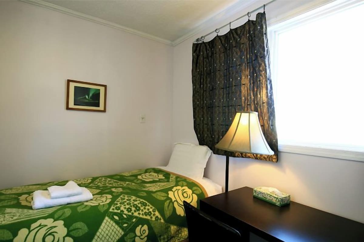 A hotel room with a green floral bedspread, a white pillow, a framed picture on the wall, a dark wooden desk with a lamp, and a window with a dark curtain.