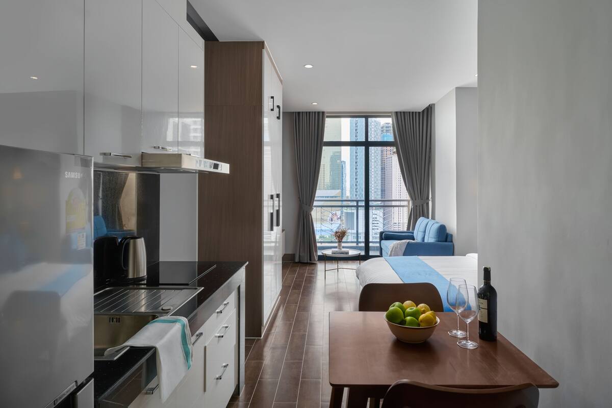 A modern hotel room with a kitchenette, a dining area with a table and chairs, a bed with a blue headboard, and a large window offering a city view.